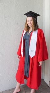 A young woman with light brown wavy hair, fair skin, and blue eyes smiles at the camera, which is in front and slightly to the left of her. She wears a black graduation cap and tassel, red pleated graduation robe, and white stole over a blue/grey long, flowy, V-neck dress.