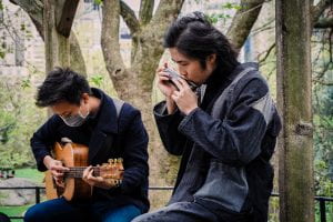 Two AAPI musicians are performing at Central Park, one playing the harmonica and the other playing the guitar.