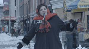 Woman wearing a red scarf around her hair and a black jacket is singing at Union Square.