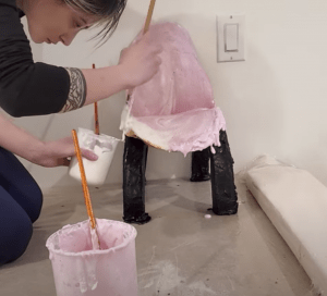 Leah Hughes builds a half-scale chair out gelatin bioplastic. Leah is using a paint bush to paint pink and white gelatin onto the seat of the chair. The chair's legs are black.