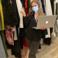 A picture of Emily standing in front of a packed clothing rack. She is holding a silver Apple laptop in one hand, the other hand is a peace sign. She is wearing black jeans, a black button-up top, and black sneakers with a red stripe. She is double-masked.