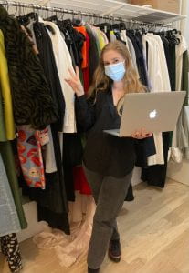 A picture of Emily standing in front of a packed clothing rack. She is holding a silver Apple laptop in one hand, the other hand is a peace sign. She is wearing black jeans, a black button-up top, and black sneakers with a red stripe. She is double-masked.