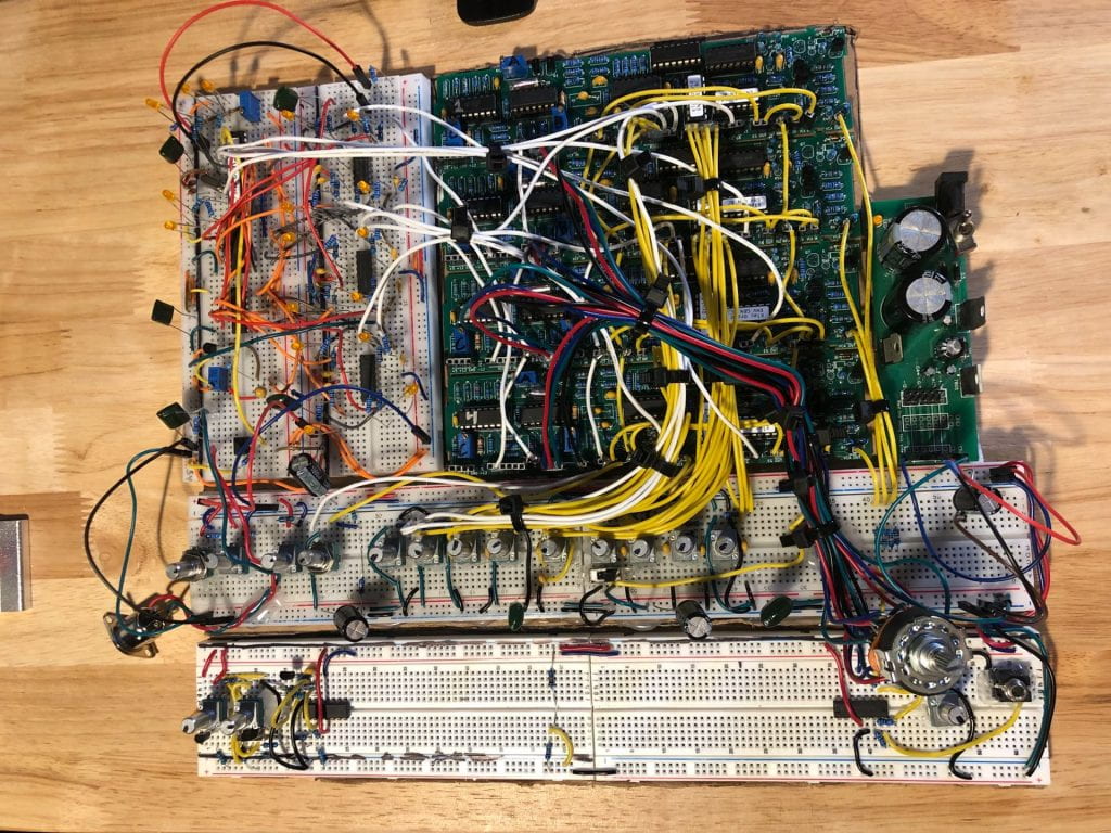 Depicted is the prototype of the synthesizer. It is a square made up of white breadboards on the bottom and left side and green circuitboards in the middle and right sides. There are many different chips and other components with many colored wires connecting everything.