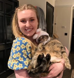 This is an image of myself with my cat, Etta in my apartment. My hair is blonde and the top half is up in two pigtails with strawberry shaped pony tails. My eyes are blue and I am wearing a blue dress. She is wearing bunny ears because it was Easter.